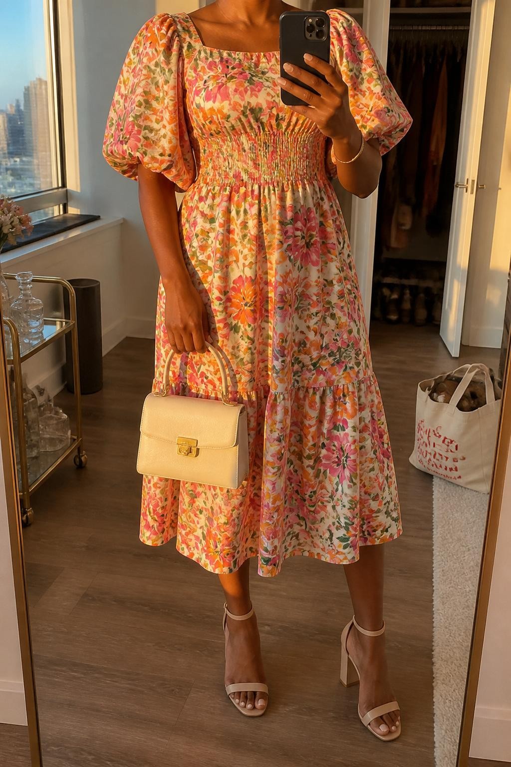 mirror selfie of a black woman wearing a bright floral cotton puff sleeve midi dress with nude block heels, a cream top-handle bag, gold stud earrings, and fresh dewy makeup, holding a phone in front of a mirror, cropped at NECK so face doesn't show, with a natural casual selfie pose, in a city apartment, evening golden hour, including small lived-in details like a tote bag on the floor and an open closet behind, latest iPhone photo quality (no watermarks on images)