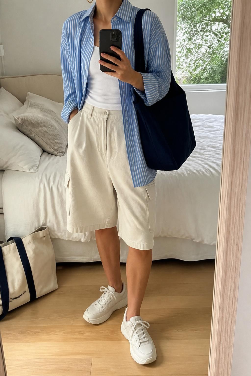 mirror selfie of a woman wearing cream baggy cargo shorts, blue and white striped oversized button-down shirt worn open over a white fitted tank, white chunky sneakers, navy canvas tote bag, and small gold hoop earrings, clearly showing the same clothing items, colors, fabrics, and accessories listed in the Outfit Pieces section, holding a phone in front of a mirror, cropped at NECK so the face does not show, with a relaxed casual selfie pose, in a cozy bedroom mirror setup, natural window light, including soft pillows on an unmade bed and a tote bag resting on the floor, latest iPhone photo quality.