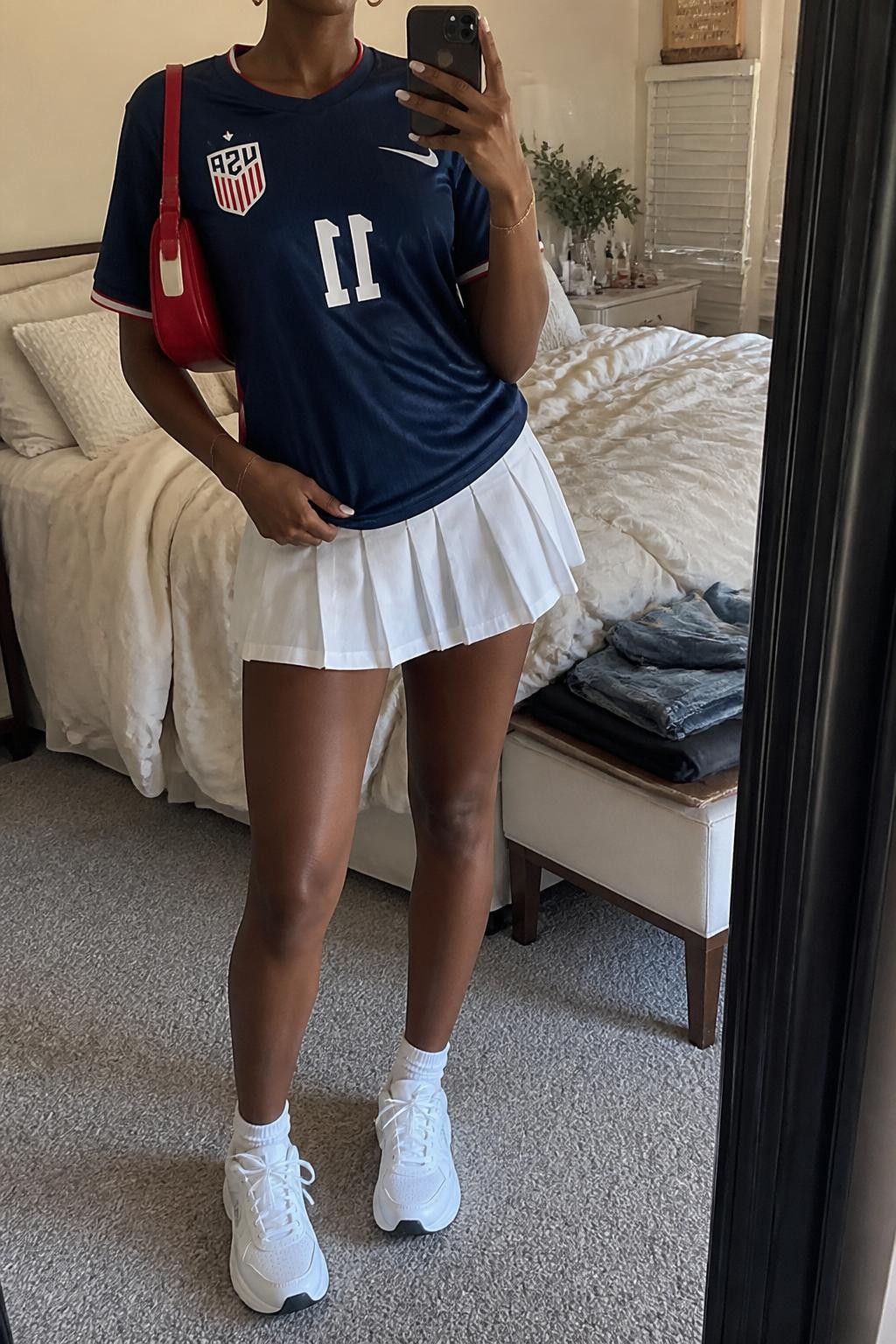 mirror selfie of a Black woman wearing a navy USA soccer jersey, white pleated tennis-style mini skirt, white chunky sneakers, red mini shoulder bag, and small gold hoop earrings, clearly showing the same clothing items, colors, fabrics, and accessories listed in the Outfit Pieces section, holding a phone in front of a mirror, cropped at NECK so the face does not show, with a relaxed casual selfie pose, in a cozy bedroom mirror setup, soft indoor lighting, including soft pillows on an unmade bed and folded denim jeans on a bench, latest iPhone photo quality.