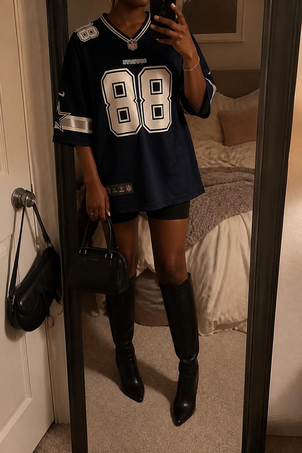 mirror selfie of a Black woman wearing an oversized navy football jersey, black biker shorts, black knee-high heeled boots, black structured mini bag, and silver hoop earrings, clearly showing the same clothing items, colors, fabrics, and accessories listed in the Outfit Pieces section, holding a phone in front of a mirror, cropped at NECK so the face does not show, with a relaxed casual selfie pose, in a cozy bedroom mirror setup, warm moody lighting, including soft pillows on an unmade bed and a handbag hanging on a door handle, latest iPhone photo quality.