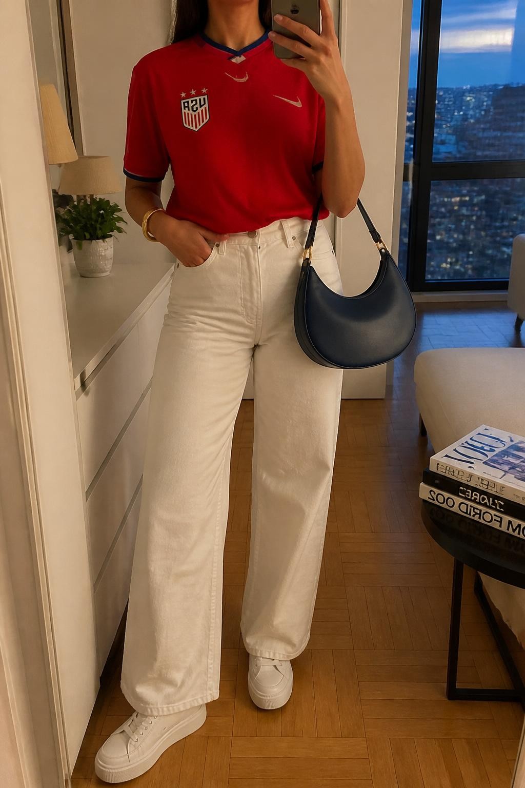 mirror selfie of a woman wearing a red USA soccer jersey tucked into white high-waisted wide-leg jeans, white platform sneakers, navy crescent shoulder bag, and gold bangle bracelet, clearly showing the same clothing items, colors, fabrics, and accessories listed in the Outfit Pieces section, holding a phone in front of a mirror, cropped at NECK so the face does not show, with a relaxed casual selfie pose, in a stylish city apartment, evening golden hour, including a small plant on a dresser and a stack of fashion magazines on a side table, latest iPhone photo quality.