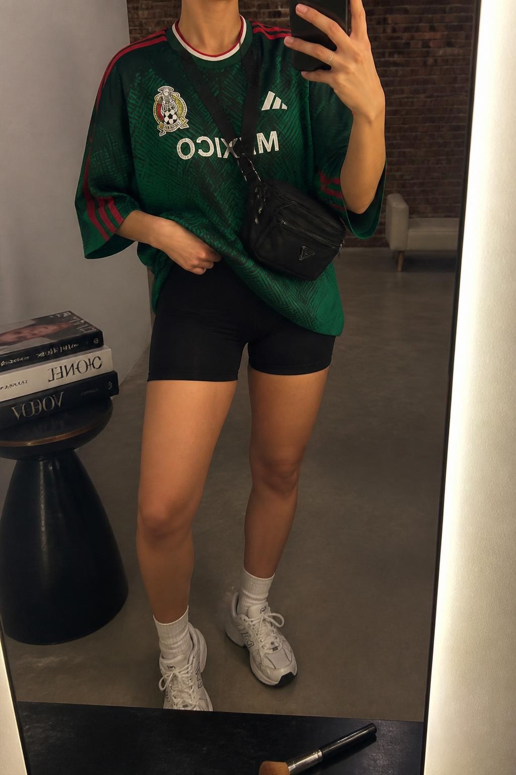 mirror selfie of a woman wearing an oversized green Mexico soccer jersey, black high-waisted biker shorts, white athletic sneakers, black nylon belt bag worn crossbody, and white crew socks, clearly showing the same clothing items, colors, fabrics, and accessories listed in the Outfit Pieces section, holding a phone in front of a mirror, cropped at NECK so the face does not show, with a relaxed casual selfie pose, in a fashion studio mirror corner, warm moody lighting, including a stack of fashion magazines on a side table and a makeup brush on the counter, latest iPhone photo quality.