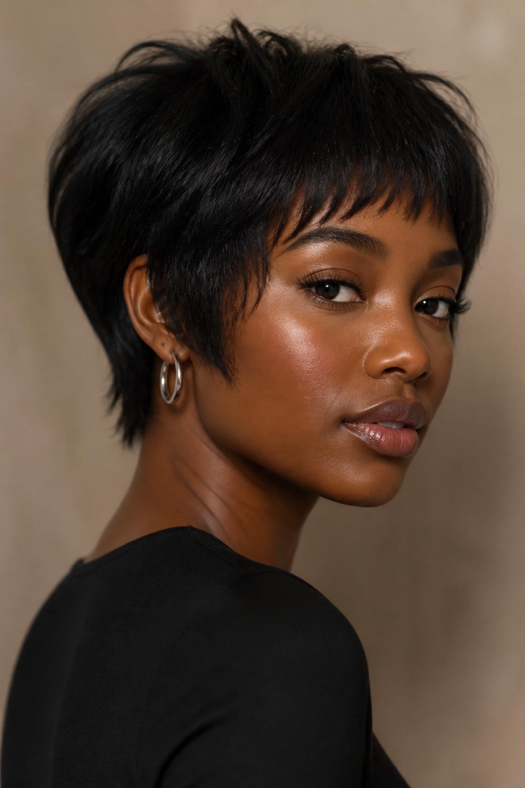A realistic portrait of a Black woman with rich dark skin wearing micro French wispy bangs with a textured pixie bob, jet black hair with soft espresso dimension, short airy fringe sitting above the brows with feathered uneven tips, longer side pieces near the temples, textured pixie-bob shape with volume through the crown, tapered nape, soft piecey layers around the ears, natural edges, silver hoop earrings, black fitted top, photographed against a warm taupe wall with soft studio lighting, realistic skin texture, natural hair detail, modern editorial short hairstyle portrait.