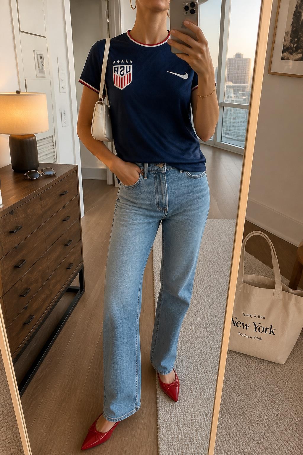 mirror selfie of a woman wearing a navy USA soccer jersey, light blue straight-leg jeans, red pointed flats, white mini shoulder bag, and gold hoop earrings, clearly showing the same clothing items, colors, fabrics, and accessories listed in the Outfit Pieces section, holding a phone in front of a mirror, cropped at NECK so the face does not show, with a relaxed casual selfie pose, in a stylish city apartment, evening golden hour, including sunglasses placed on a dresser and a cream tote bag resting on the floor, latest iPhone photo quality.