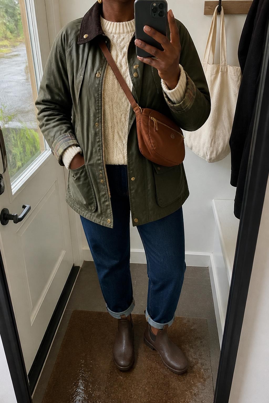 mirror selfie of a Black woman wearing the exact outfit described above, clearly showing an olive waxed barn jacket layered over a cream cable knit sweater, dark blue cuffed jeans, brown waterproof ankle boots, a cognac nylon crossbody bag, and gold stud earrings, holding a phone in front of a mirror, cropped at NECK so the face does not show, with a relaxed casual selfie pose, in a small mudroom-style entry mirror, natural rainy daylight, with a tote bag hanging on a hook and a damp doormat near the boots, latest iPhone photo quality.