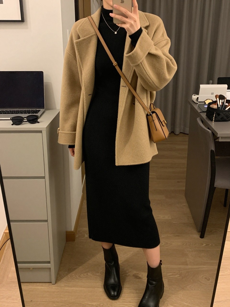 mirror selfie of a woman wearing a tan wool-blend shacket over a black fitted mock-neck top, black knit midi skirt, black ankle boots, and a tan crossbody bag, clearly showing the same clothing items, colors, fabrics, and accessories listed in the Outfit Pieces section, holding a phone in front of a mirror, cropped at NECK so the face does not show, with a relaxed casual selfie pose, in a stylish city apartment, warm moody lighting, including sunglasses placed on a dresser and a makeup brush or cosmetic bag on the counter, latest iPhone photo quality.