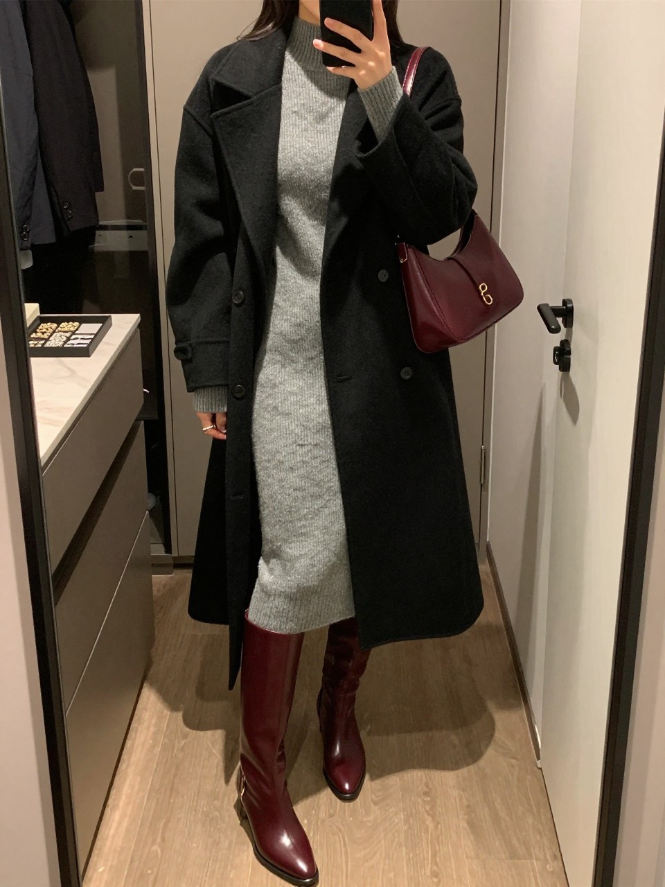 mirror selfie of a woman wearing a gray ribbed midi sweater dress, burgundy knee-high leather boots, long black wool coat, slim gold hoops, and a burgundy shoulder bag, clearly showing the same clothing items, colors, fabrics, and accessories listed in the Outfit Pieces section, holding a phone in front of a mirror, cropped at NECK so the face does not show, with a relaxed casual selfie pose, in a walk-in closet mirror, warm moody lighting, including a jewelry tray on the vanity and a handbag hanging on a door handle, latest iPhone photo quality.