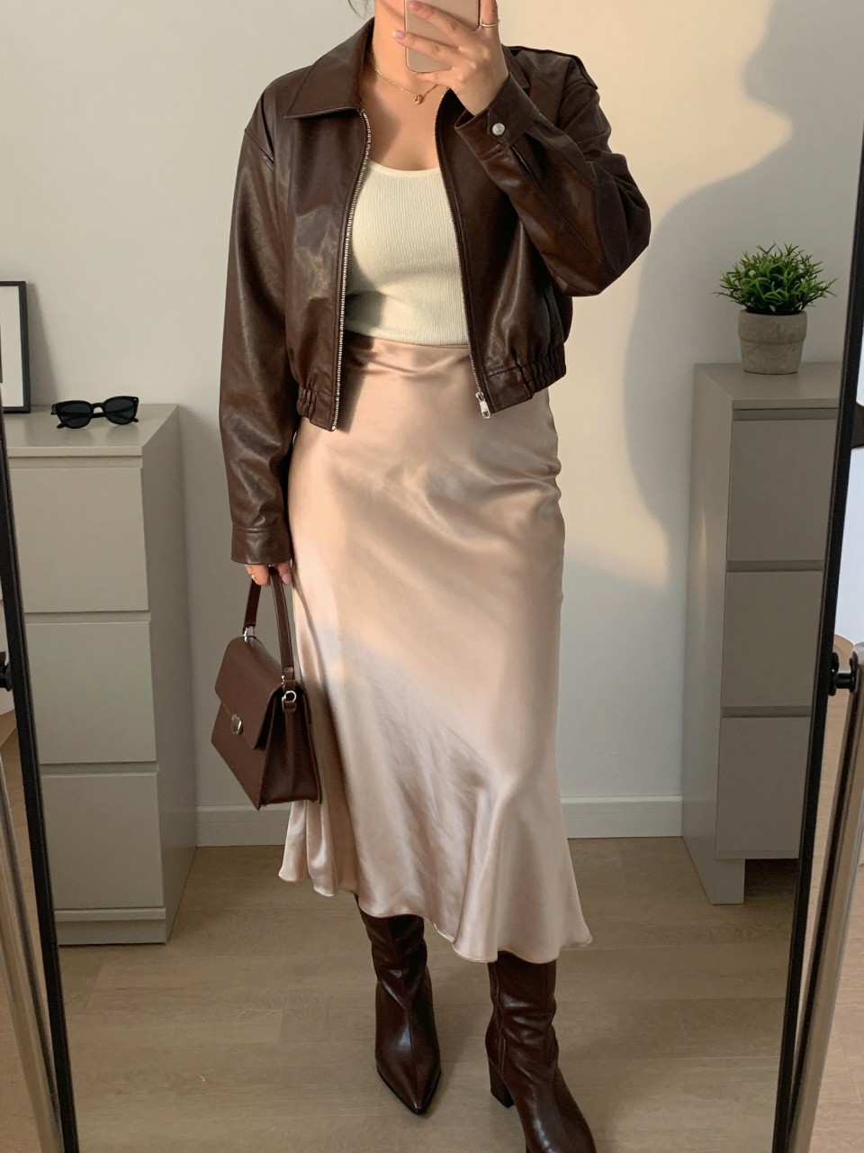 mirror selfie of a curvy woman wearing a chocolate brown leather jacket over a fitted ivory knit top, champagne satin midi slip skirt, brown pointed ankle boots, and a small chocolate brown handbag, clearly showing the same clothing items, colors, fabrics, and accessories listed in the Outfit Pieces section, holding a phone in front of a mirror, cropped at NECK so the face does not show, with a relaxed casual selfie pose, in a stylish city apartment, evening golden hour, including sunglasses placed on a dresser and a small plant on a dresser, latest iPhone photo quality.