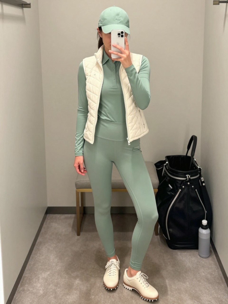 Mirror selfie of a woman wearing a slim cream quilted golf vest over a fitted sage green long-sleeve polo, matching sage high-waisted golf leggings with side pockets, cream spikeless golf shoes, and a structured sage baseball cap, clearly showing the exact clothing items, colors, fabrics, and accessories listed, holding a latest-model iPhone in front of a mirror, cropped at the neck so the face does not show, relaxed casual selfie pose, in a minimal dressing room with soft indoor lighting, a golf tote leaning against a bench and a water bottle on the floor, natural uncluttered composition, latest iPhone photo quality.