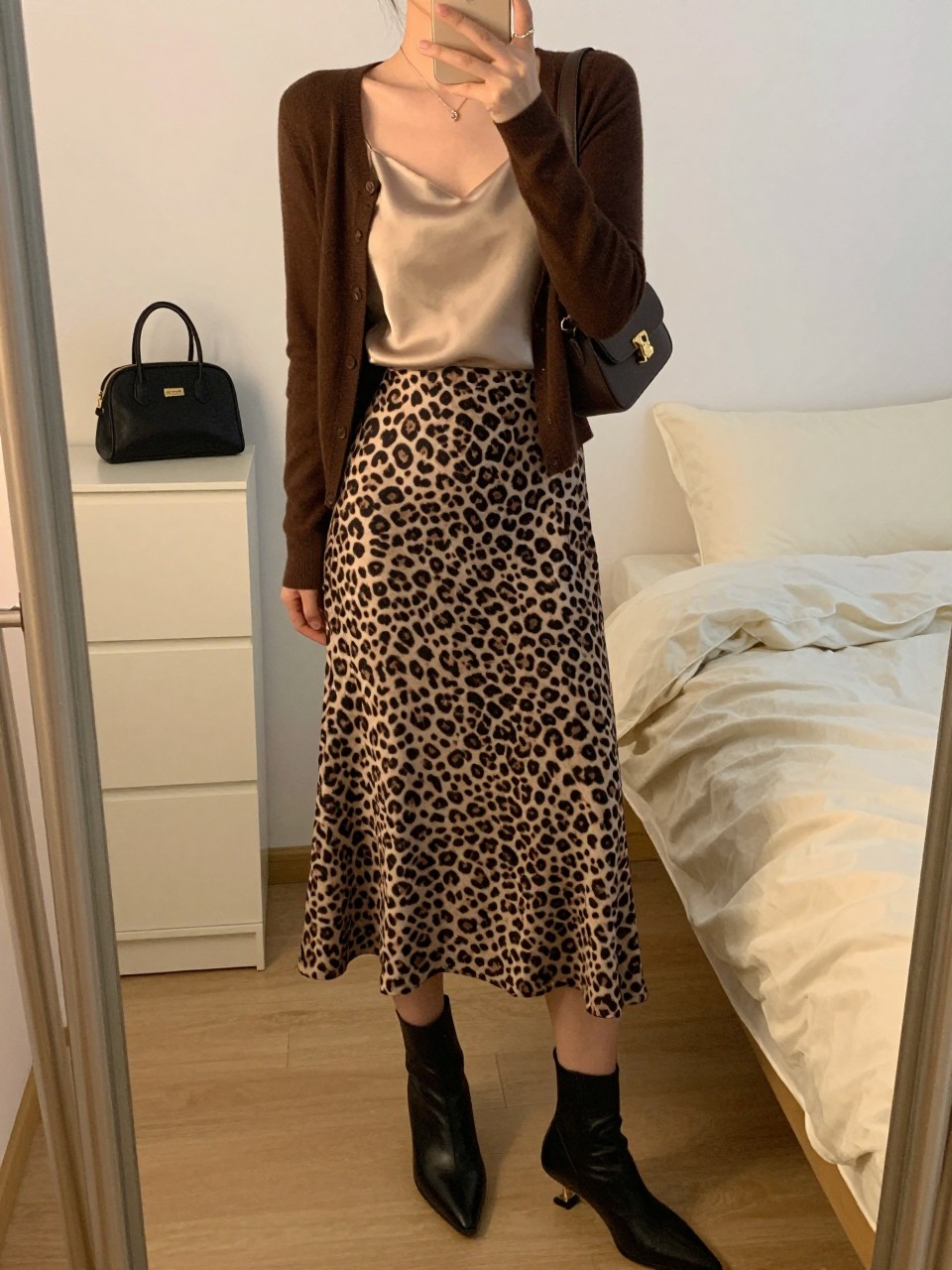 mirror selfie of a woman wearing a chocolate brown fitted cardigan, beige silk camisole, subtle brown and black leopard-print midi skirt, black kitten-heel ankle boots, and a small dark brown shoulder bag, clearly showing the same clothing items, colors, fabrics, and accessories listed in the Outfit Pieces section, holding a phone in front of a mirror, cropped at NECK so the face does not show, with a relaxed casual selfie pose, in a feminine-themed bedroom, evening golden hour, including a cosmetic bag on the dresser and soft pillows on an unmade bed, latest iPhone photo quality.