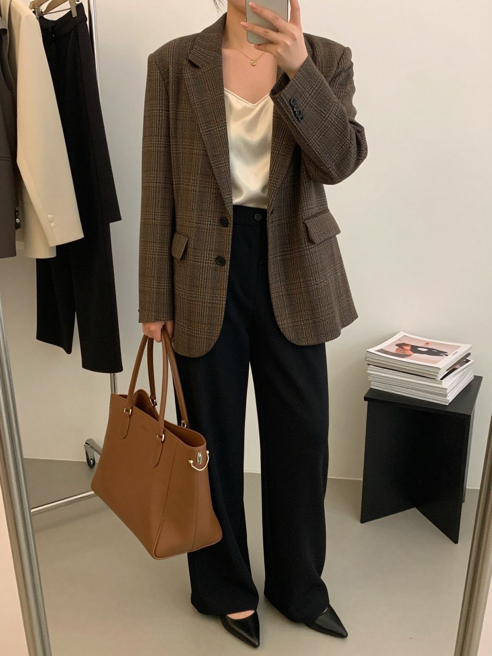 mirror selfie of a curvy woman wearing a brown plaid blazer over a cream silk camisole, black high-waisted wide-leg trousers, black pointed pumps, and a structured brown work tote, clearly showing the same clothing items, colors, fabrics, and accessories listed in the Outfit Pieces section, holding a phone in front of a mirror, cropped at NECK so the face does not show, with a relaxed casual selfie pose, in a fashion studio mirror corner, soft indoor lighting, including a clothing rack with garments behind and a stack of fashion magazines on a side table, latest iPhone photo quality.