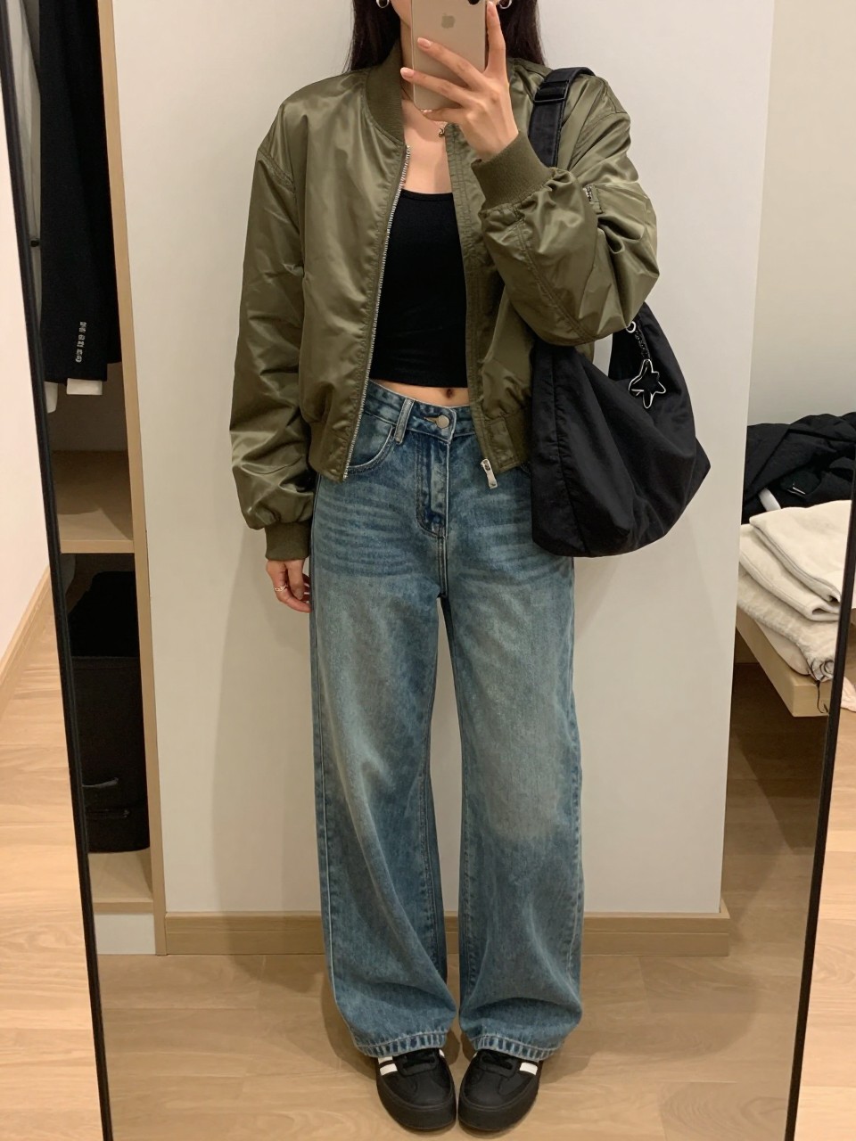 mirror selfie of a woman wearing an olive satin bomber jacket, fitted black cropped tank, medium-wash wide-leg denim jeans, black platform sneakers, silver hoop earrings, and a black nylon shoulder bag, clearly showing the same clothing items, colors, fabrics, and accessories listed in the Outfit Pieces section, holding a phone in front of a mirror, cropped at NECK so the face does not show, with a relaxed casual selfie pose, in a walk-in closet mirror, soft indoor lighting, including an open closet with clothes visible and a folded blanket on a chair, latest iPhone photo quality.