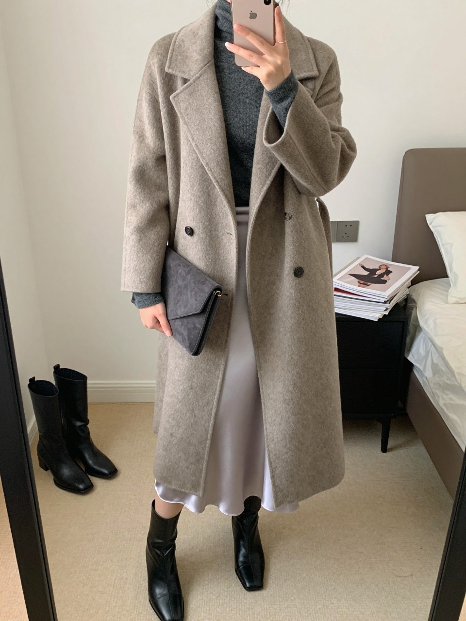 Mirror selfie of a woman wearing a long belted taupe wool wrap coat over a fitted charcoal gray cashmere turtleneck, a silver satin midi skirt, black leather square-toe ankle boots, and a charcoal gray suede clutch, clearly showing the exact clothing items, colors, textures, and accessories listed, holding a latest-model iPhone in front of a mirror, cropped at the neck so the face does not show, relaxed casual selfie pose, in a feminine-themed bedroom with soft indoor lighting, a pair of boots on the floor nearby and a stack of fashion magazines on a side table, natural unstaged composition, latest iPhone photo quality.