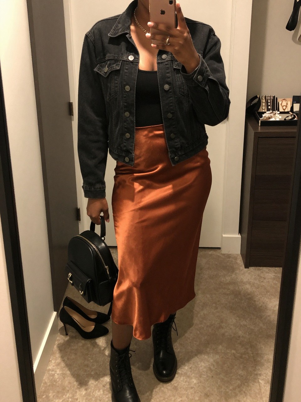 mirror selfie of a Black woman wearing the exact outfit described above, clearly showing a black denim jacket, fitted black tank, rust satin slip skirt, black combat boots, and a small black backpack purse, holding a phone in front of a mirror, cropped at NECK so the face does not show, with a relaxed casual selfie pose, in a walk-in closet mirror, warm moody lighting, with a pair of heels on the floor nearby and a jewelry tray on the vanity, latest iPhone photo quality.