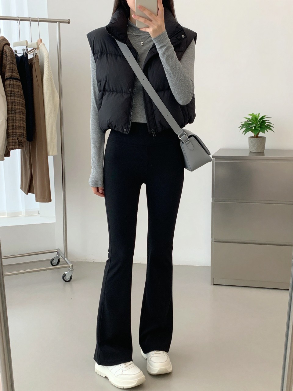 mirror selfie of a woman wearing a cropped black puffer vest over a gray fitted long-sleeve top, black flared leggings, white chunky sneakers, and a gray crossbody sling bag, clearly showing the same clothing items, colors, fabrics, and accessories listed in the Outfit Pieces section, holding a phone in front of a mirror, cropped at NECK so the face does not show, with a relaxed casual selfie pose, in a minimal dressing room, natural window light, including a clothing rack with garments behind and a small plant on a dresser, latest iPhone photo quality.