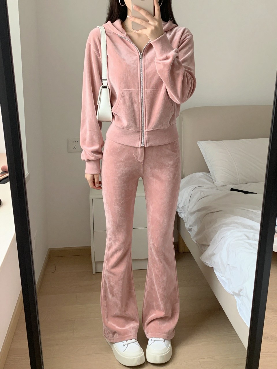 mirror selfie of a woman wearing a dusty pink velour zip-up hoodie, matching dusty pink velour flared pants, white platform sneakers, silver hoop earrings, and a tiny white shoulder bag, clearly showing the same clothing items, colors, fabrics, and accessories listed in the Outfit Pieces section, holding a phone in front of a mirror, cropped at NECK so the face does not show, with a relaxed casual selfie pose, in a feminine-themed bedroom, soft indoor lighting, including soft pillows on an unmade bed and a makeup brush on the counter, latest iPhone photo quality.