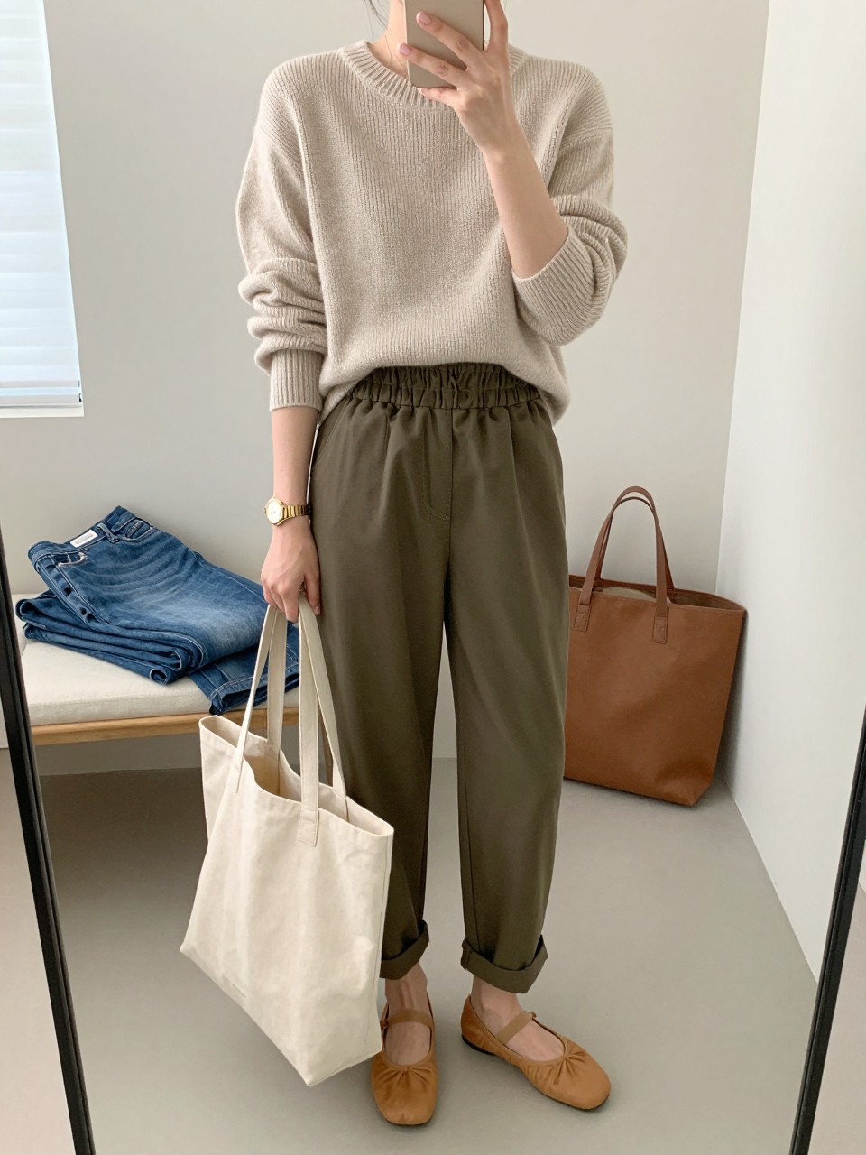 mirror selfie of a woman wearing an oatmeal knit pullover sweater, olive green paperbag waist pants, tan ballet flats, gold watch, and a beige canvas teacher tote, clearly showing the same clothing items, colors, fabrics, and accessories listed in the Outfit Pieces section, holding a phone in front of a mirror, cropped at NECK so the face does not show, with a relaxed casual selfie pose, in a minimal dressing room, natural window light, including folded denim jeans on a bench and a tote bag resting on the floor, latest iPhone photo quality.
