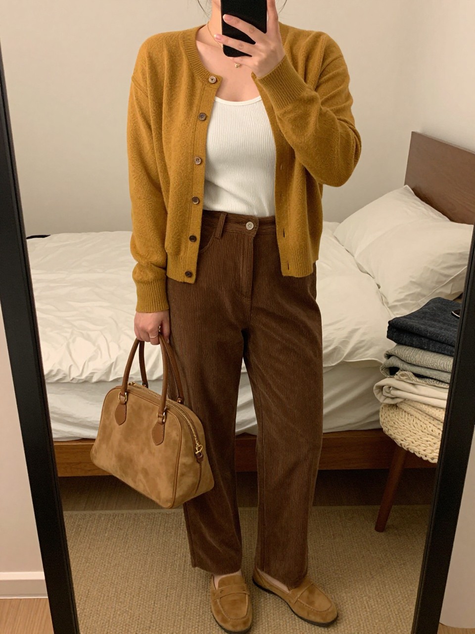 mirror selfie of a curvy woman wearing a mustard button-front cardigan over a white ribbed tank top, brown high-waisted corduroy pants, tan loafers, and a tan suede handbag, clearly showing the same clothing items, colors, fabrics, and accessories listed in the Outfit Pieces section, holding a phone in front of a mirror, cropped at NECK so the face does not show, with a relaxed casual selfie pose, in a cozy bedroom mirror setup, warm moody lighting, including soft pillows on an unmade bed and a folded blanket on a chair, latest iPhone photo quality.