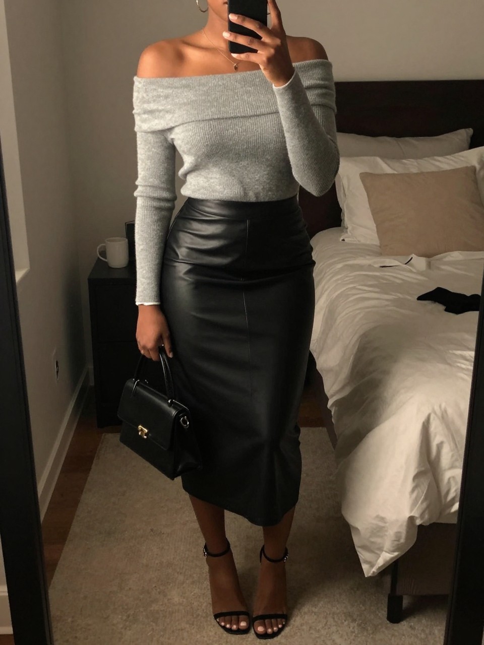mirror selfie of a Black woman wearing the exact outfit described above, clearly showing a soft gray off-shoulder knit top, black leather midi skirt, black ankle-strap heels, silver bracelet, and a black mini handbag, holding a phone in front of a mirror, cropped at NECK so the face does not show, with a relaxed casual selfie pose, in a feminine-themed bedroom, warm moody lighting, with a coffee mug on a bedside table and soft pillows on an unmade bed, latest iPhone photo quality.
