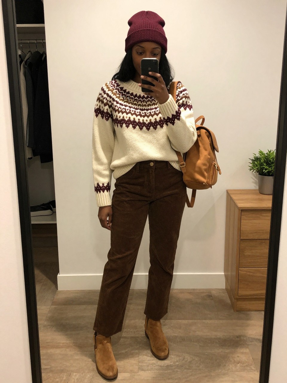 mirror selfie of a Black woman wearing the exact outfit described above, clearly showing a cream Fair Isle sweater with brown and burgundy pattern, brown corduroy straight-leg pants, tan suede ankle boots, burgundy beanie, and a small tan backpack, holding a phone in front of a mirror, cropped at NECK so the face does not show, with a relaxed casual selfie pose, in a minimal dressing room, warm moody lighting, with an open closet with clothes visible and a small plant on a dresser, latest iPhone photo quality.