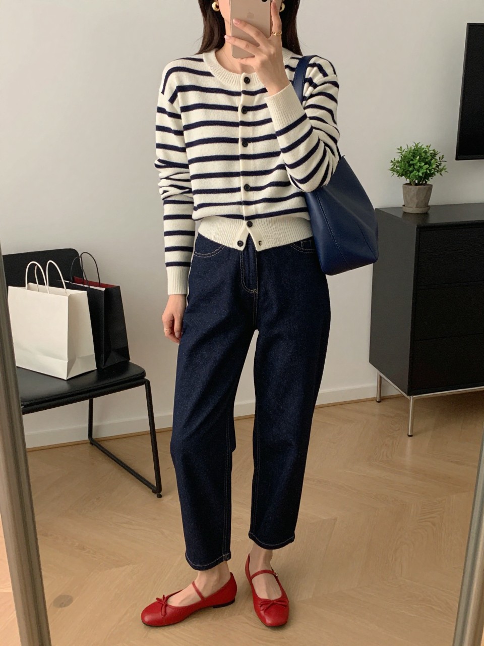 mirror selfie of a woman wearing the exact outfit described above, clearly showing dark indigo barrel jeans, a navy and cream striped button-front cardigan, red ballet flats, a navy leather shoulder bag, and small gold huggie earrings, holding a phone in front of a mirror, cropped at NECK so the face does not show, with a relaxed casual selfie pose, in a stylish city apartment, soft indoor lighting, with shopping bags beside a chair and a small plant on a dresser, latest iPhone photo quality.