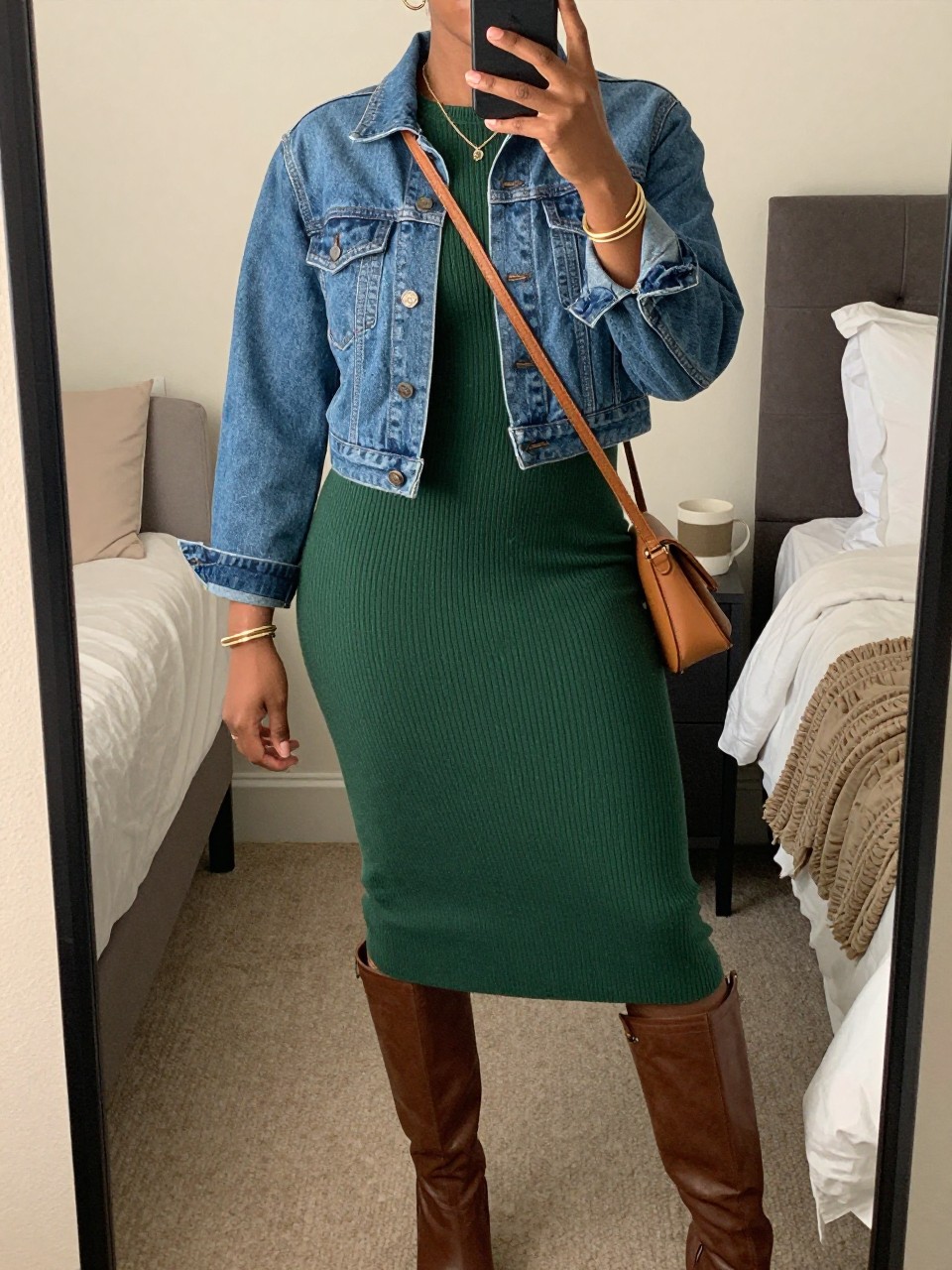 mirror selfie of a Black woman wearing the exact outfit described above, clearly showing a cropped medium-wash denim jacket layered over a forest green ribbed sweater midi dress, brown knee-high boots, a tan crossbody bag, and gold bangles, holding a phone in front of a mirror, cropped at NECK so the face does not show, with a relaxed casual selfie pose, in a cozy bedroom mirror setup, soft indoor lighting, with a coffee mug on a bedside table and a folded blanket on a chair, latest iPhone photo quality.