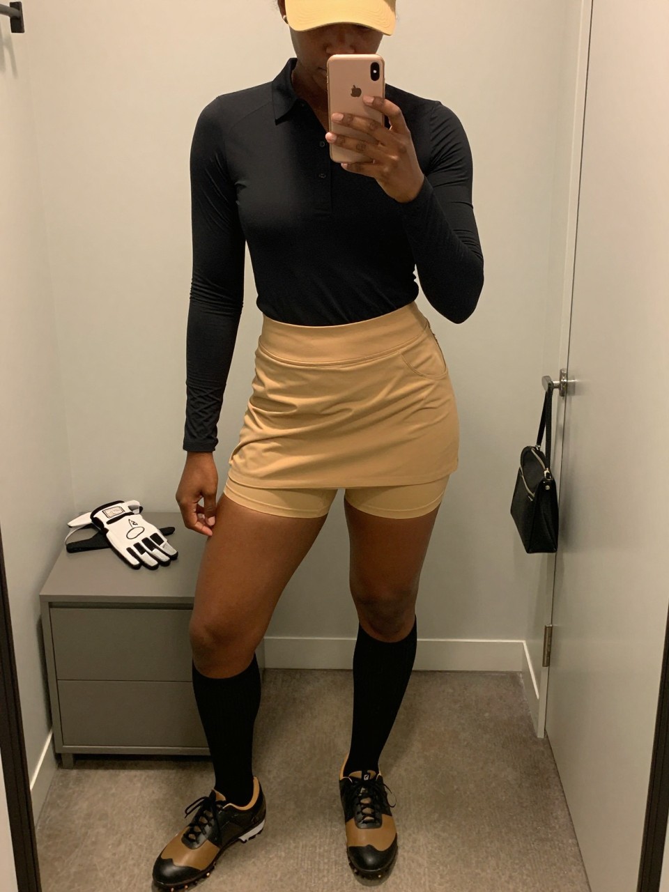 Mirror selfie of a Black woman with deep brown skin wearing a fitted black long-sleeve performance golf polo, a camel asymmetrical wrap-front golf skort with hidden built-in shorts, black knee-high ribbed socks, black-and-camel spikeless golf shoes, and a camel visor, clearly showing the exact clothing items, colors, fabrics, and accessories listed, holding a latest-model iPhone in front of a mirror, cropped at the neck so the face does not show, relaxed casual selfie pose, in a minimal dressing room with warm moody lighting, a golf glove resting on a dresser and a small handbag hanging on a door handle, natural uncluttered setting, latest iPhone photo quality.