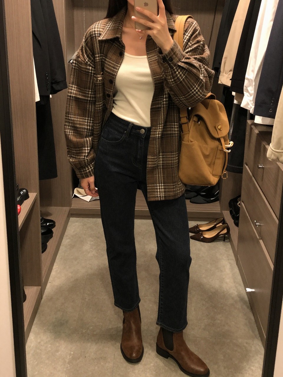 mirror selfie of a woman wearing a brown plaid oversized shacket over a cream fitted tank top, dark wash straight-leg jeans, brown ankle boots, and a tan backpack purse, clearly showing the same clothing items, colors, fabrics, and accessories listed in the Outfit Pieces section, holding a phone in front of a mirror, cropped at NECK so the face does not show, with a relaxed casual selfie pose, in a walk-in closet mirror, warm moody lighting, including open closet with clothes visible and a pair of heels on the floor nearby, latest iPhone photo quality.