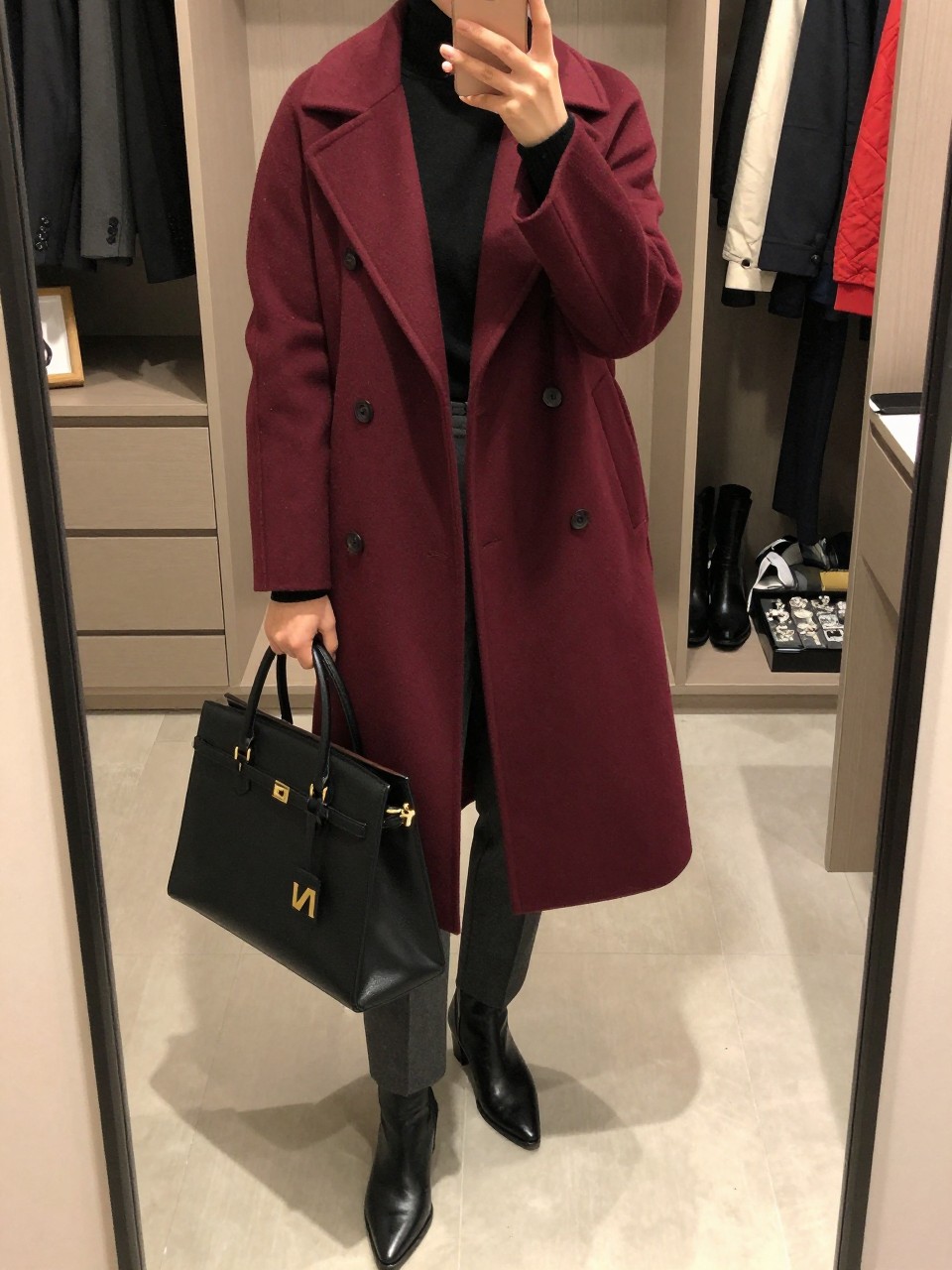 mirror selfie of a woman wearing a burgundy wool coat over a black fine-knit turtleneck, dark gray tailored trousers, black pointed leather boots, and a black structured work bag, clearly showing the same clothing items, colors, fabrics, and accessories listed in the Outfit Pieces section, holding a phone in front of a mirror, cropped at NECK so the face does not show, with a relaxed casual selfie pose, in a walk-in closet mirror, soft indoor lighting, including open closet with clothes visible and a jewelry tray on the vanity, latest iPhone photo quality.