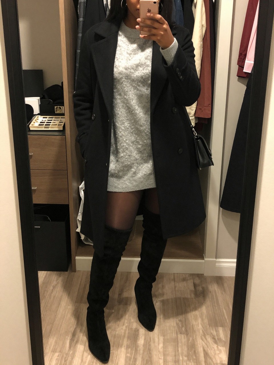 mirror selfie of a Black woman wearing the exact outfit described above, clearly showing black over-the-knee suede boots, a gray oversized sweater dress, sheer black tights, a black wool coat, a black mini shoulder bag, and silver hoop earrings, holding a phone in front of a mirror, cropped at NECK so the face does not show, with a relaxed casual selfie pose, in a walk-in closet mirror, warm moody lighting, with an open closet with clothes visible and a jewelry tray on the vanity, latest iPhone photo quality.