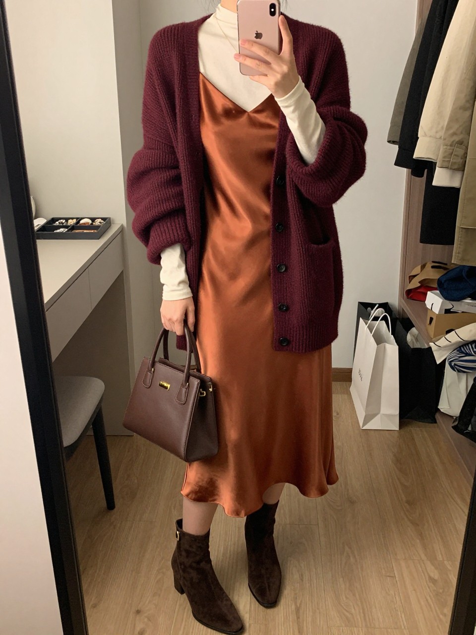 Mirror selfie of a woman wearing a chunky burgundy cardigan over a copper satin midi slip dress layered above a fitted cream long-sleeve top, dark brown suede ankle boots, and a small structured chocolate brown handbag, clearly showing the exact clothing items, colors, fabrics, and accessories listed, holding a latest-model iPhone in front of a mirror, cropped at the neck so the face does not show, relaxed casual selfie pose, in a walk-in closet mirror with evening golden-hour lighting, a jewelry tray on the vanity and shopping bags beside a chair, natural unstaged composition, latest iPhone photo quality.