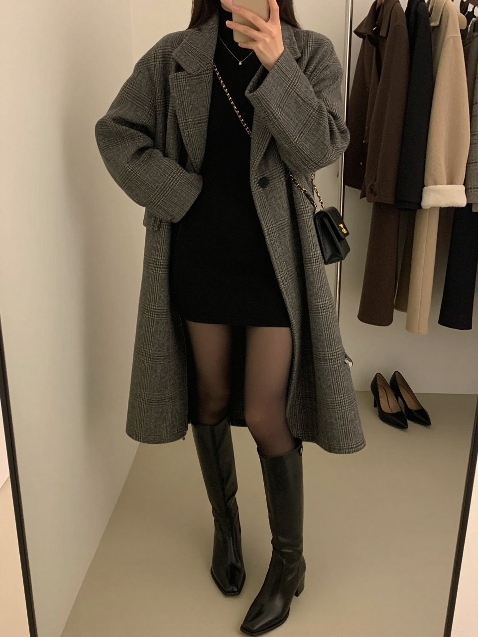 mirror selfie of a woman wearing a gray plaid wool coat, fitted black long-sleeve mini dress, sheer black tights, black knee-high leather boots, and a small black chain-strap bag, clearly showing the same clothing items, colors, fabrics, and accessories listed in the Outfit Pieces section, holding a phone in front of a mirror, cropped at NECK so the face does not show, with a relaxed casual selfie pose, in a fashion studio mirror corner, warm moody lighting, including a clothing rack with garments behind and a pair of heels on the floor nearby, latest iPhone photo quality.