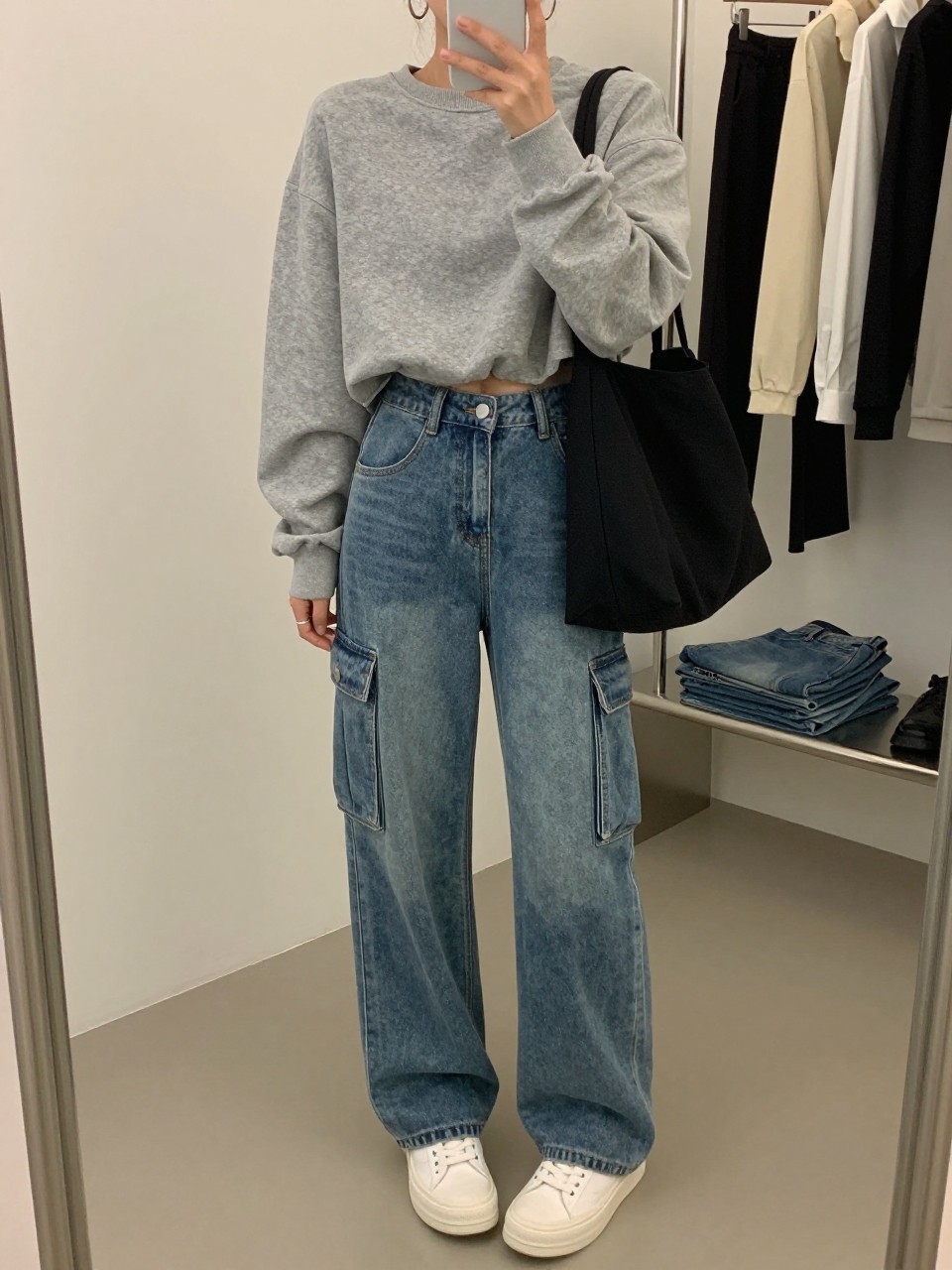 mirror selfie of a woman wearing a gray cropped sweatshirt, medium wash denim cargo jeans, white platform sneakers, silver hoop earrings, and a black canvas tote bag, clearly showing the same clothing items, colors, fabrics, and accessories listed in the Outfit Pieces section, holding a phone in front of a mirror, cropped at NECK so the face does not show, with a relaxed casual selfie pose, in a fashion studio mirror corner, soft indoor lighting, including a clothing rack with garments behind and folded denim jeans on a bench, latest iPhone photo quality.