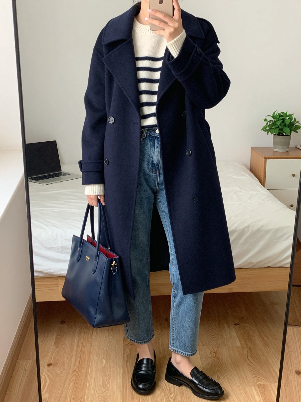 mirror selfie of a woman wearing a navy wool peacoat over an ivory and navy striped cashmere sweater, medium wash straight jeans, black loafers, and a navy leather tote, clearly showing the same clothing items, colors, fabrics, and accessories listed in the Outfit Pieces section, holding a phone in front of a mirror, cropped at NECK so the face does not show, with a relaxed casual selfie pose, in a cozy bedroom mirror setup, natural window light, including a laptop on the bed and a small plant on a dresser, latest iPhone photo quality.