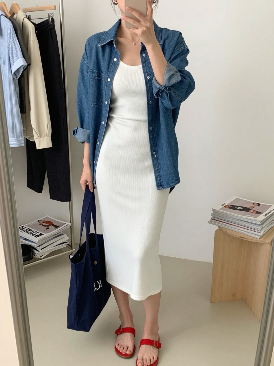 mirror selfie of a woman wearing a white ribbed fitted midi tank dress layered under an open medium blue denim shirt, red flat sandals, navy canvas tote bag, and small silver hoop earrings, clearly showing the same clothing items, colors, fabrics, and accessories listed in the Outfit Pieces section, holding a phone in front of a mirror, cropped at NECK so the face does not show, with a relaxed casual selfie pose, in a fashion studio mirror corner, natural window light, including a clothing rack with garments behind and a stack of fashion magazines on a side table, latest iPhone photo quality.
