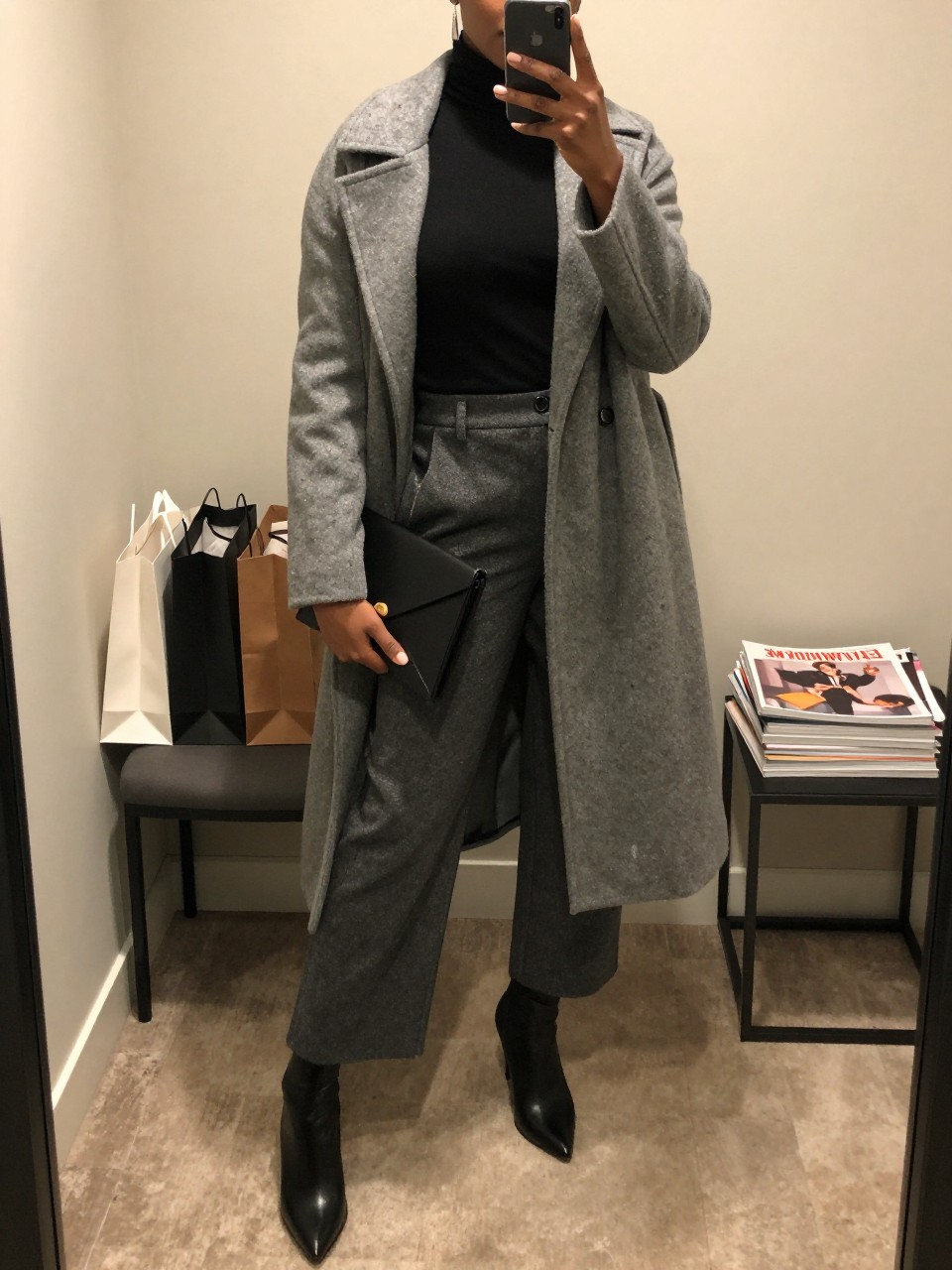 mirror selfie of a Black woman wearing the exact outfit described above, clearly showing black stiletto-heel boots, charcoal wide-leg trousers, a fitted black turtleneck, a gray long coat worn open, a black envelope clutch, and silver drop earrings, holding a phone in front of a mirror, cropped at NECK so the face does not show, with a relaxed casual selfie pose, in a minimal dressing room, warm moody lighting, with shopping bags beside a chair and a stack of fashion magazines on a side table, latest iPhone photo quality.