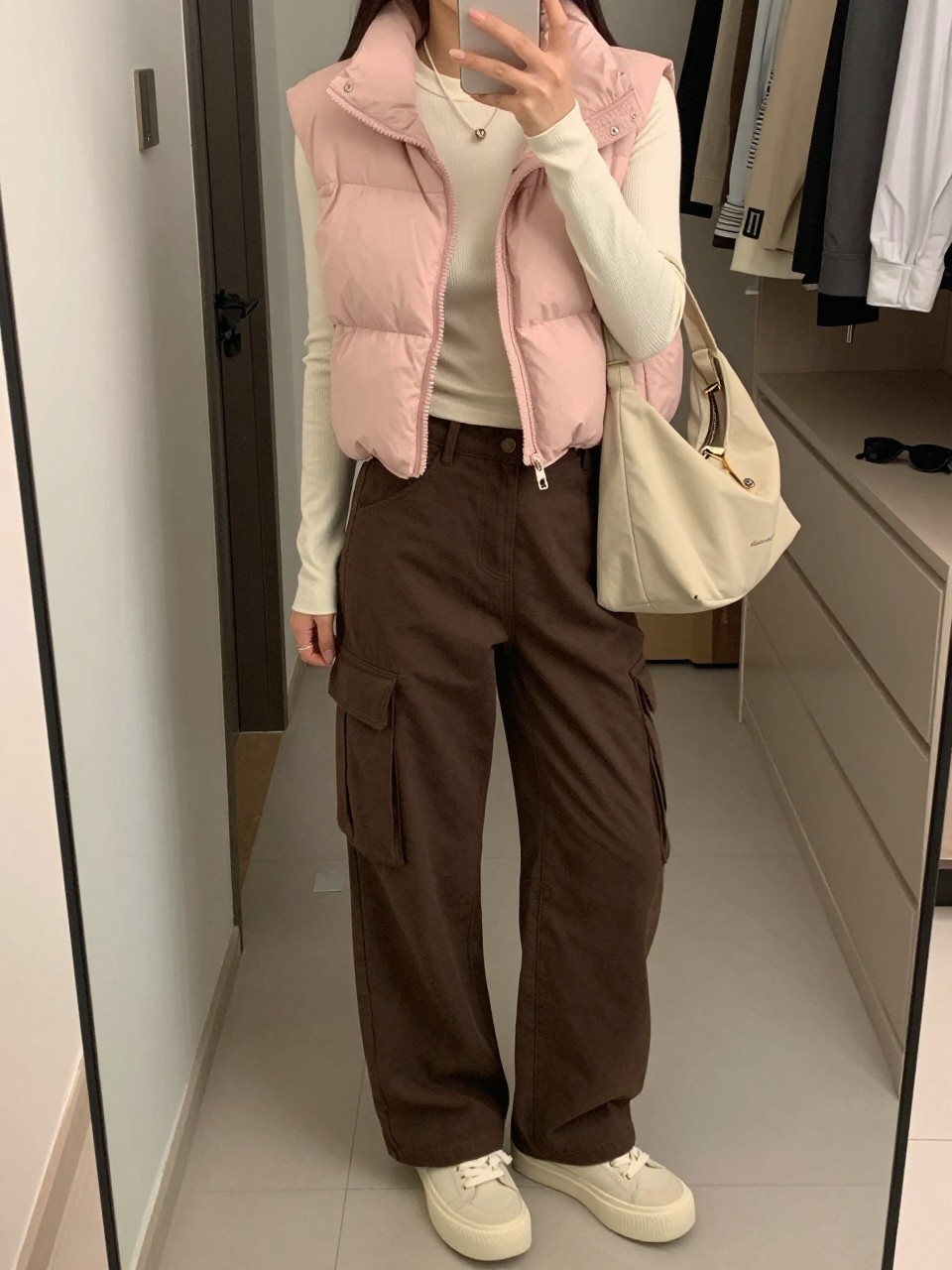 mirror selfie of a woman wearing a blush pink cropped puffer vest over a fitted ivory ribbed long-sleeve top, chocolate brown cargo pants, cream platform sneakers, and a beige nylon shoulder bag, clearly showing the same clothing items, colors, fabrics, and accessories listed in the Outfit Pieces section, holding a phone in front of a mirror, cropped at NECK so the face does not show, with a relaxed casual selfie pose, in a walk-in closet mirror, soft indoor lighting, including an open closet with clothes visible and sunglasses placed on a dresser, latest iPhone photo quality.