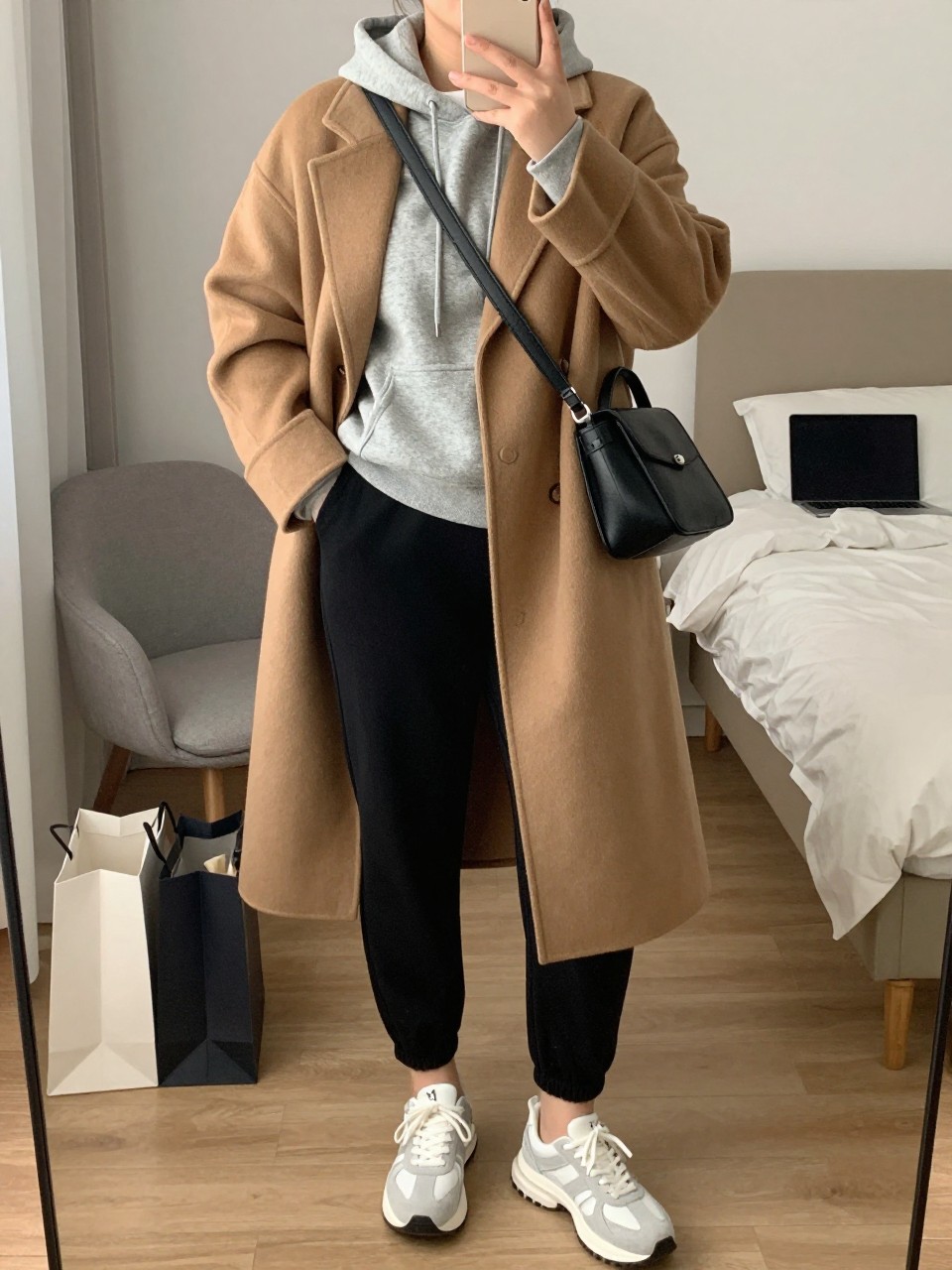 mirror selfie of a plus size woman wearing the exact outfit described above, a classic camel relaxed wool coat layered over a soft heather gray fleece hoodie, black ponte tailored jogger pants, white and gray chunky sneakers, and black leather crossbody bag, clearly showing the same clothing items, colors, fabrics, and accessories listed in the Outfit Pieces section, holding a phone in front of a mirror, cropped at NECK so the face does not show, with a relaxed casual selfie pose, in a stylish city apartment, soft indoor lighting, including shopping bags beside a chair and a laptop or tablet on the bed, latest iPhone photo quality.