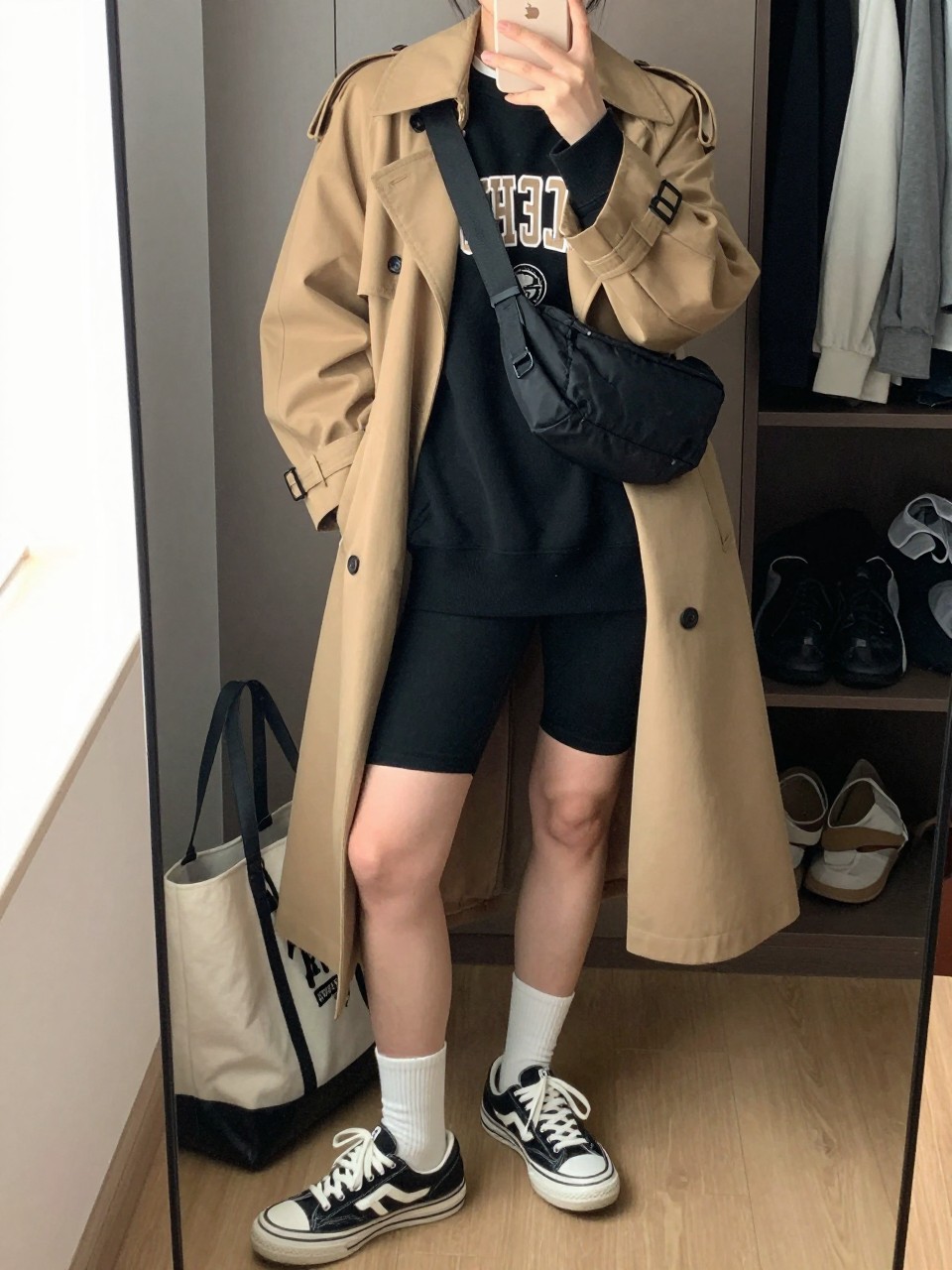 mirror selfie of a woman wearing a camel trench coat over a black oversized college sweatshirt, black bike shorts, white crew socks, black and white sneakers, and a black nylon belt bag worn crossbody, clearly showing the same clothing items, colors, fabrics, and accessories listed in the Outfit Pieces section, holding a phone in front of a mirror, cropped at NECK so the face does not show, with a relaxed casual selfie pose, in a walk-in closet mirror, natural window light, including a tote bag resting on the floor and open closet with clothes visible, latest iPhone photo quality.
