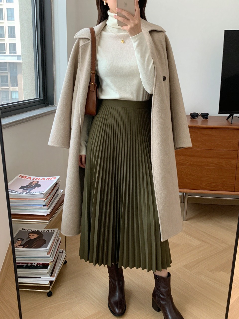 mirror selfie of a woman wearing the exact outfit described above, clearly showing an olive green pleated maxi skirt, fitted ivory turtleneck sweater, dark brown ankle boots, beige belted coat worn open, brown leather shoulder bag, and pearl stud earrings, holding a phone in front of a mirror, cropped at NECK so the face does not show, with a relaxed casual selfie pose, in a stylish city apartment, natural window light, with a stack of fashion magazines on a side table and sunglasses placed on a dresser, latest iPhone photo quality.