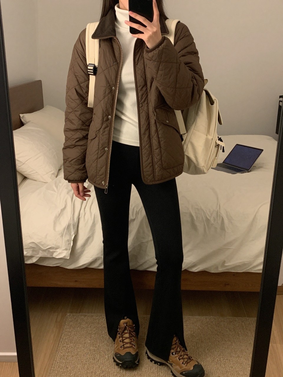 mirror selfie of a woman wearing the exact outfit described above, clearly showing a brown quilted jacket, ivory fitted hiking turtleneck, black flare leggings, brown trail boots, and a cream canvas mini backpack, holding a phone in front of a mirror, cropped at NECK so the face does not show, with a relaxed casual selfie pose, in a cozy bedroom mirror setup, warm moody lighting, with soft pillows on an unmade bed and a laptop or tablet on the bed, latest iPhone photo quality.