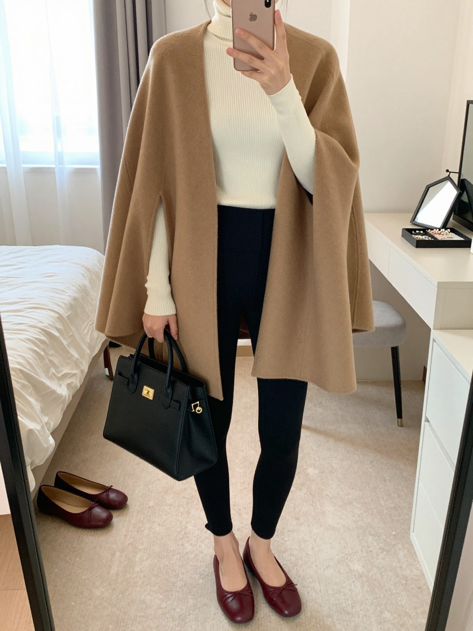 Mirror selfie of a woman wearing a structured camel wool cape over a fitted cream ribbed turtleneck, black high-waisted stirrup leggings, dark burgundy leather ballet flats, and a structured black top-handle bag, clearly showing the exact clothing items, colors, fabrics, and accessories listed, holding a latest-model iPhone in front of a mirror, cropped at the neck so the face does not show, relaxed casual selfie pose, in a feminine-themed bedroom with natural window light, a pair of flats on the floor nearby and a jewelry tray on the vanity, realistic casual composition, latest iPhone photo quality.