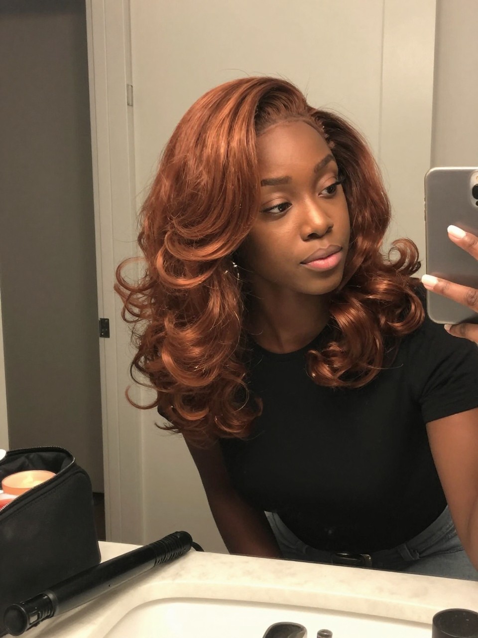 Image prompt: Photo of a Black woman wearing shoulder-length copper wand curls with a deep side part, glossy curved ends, and full side-swept volume, candid mirror selfie taken slightly from the side, bathroom vanity mirror setting, soft indoor lighting, curling wand cooling on the counter beside a makeup bag, one statement earring visible through the curls, realistic fall dinner getting-ready moment, latest iPhone photo quality.