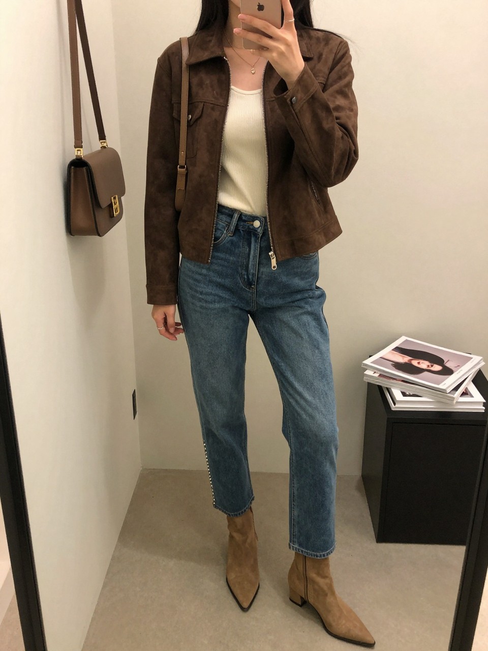 mirror selfie of a woman wearing a chocolate brown suede jacket, fitted ivory ribbed top, medium-wash bootcut jeans with rhinestone side details, pointed tan ankle boots, and a small brown shoulder bag, clearly showing the same clothing items, colors, fabrics, and accessories listed in the Outfit Pieces section, holding a phone in front of a mirror, cropped at NECK so the face does not show, with a relaxed casual selfie pose, in a minimal dressing room, warm moody lighting, including a handbag hanging on a door handle and a stack of fashion magazines on a side table, latest iPhone photo quality.
