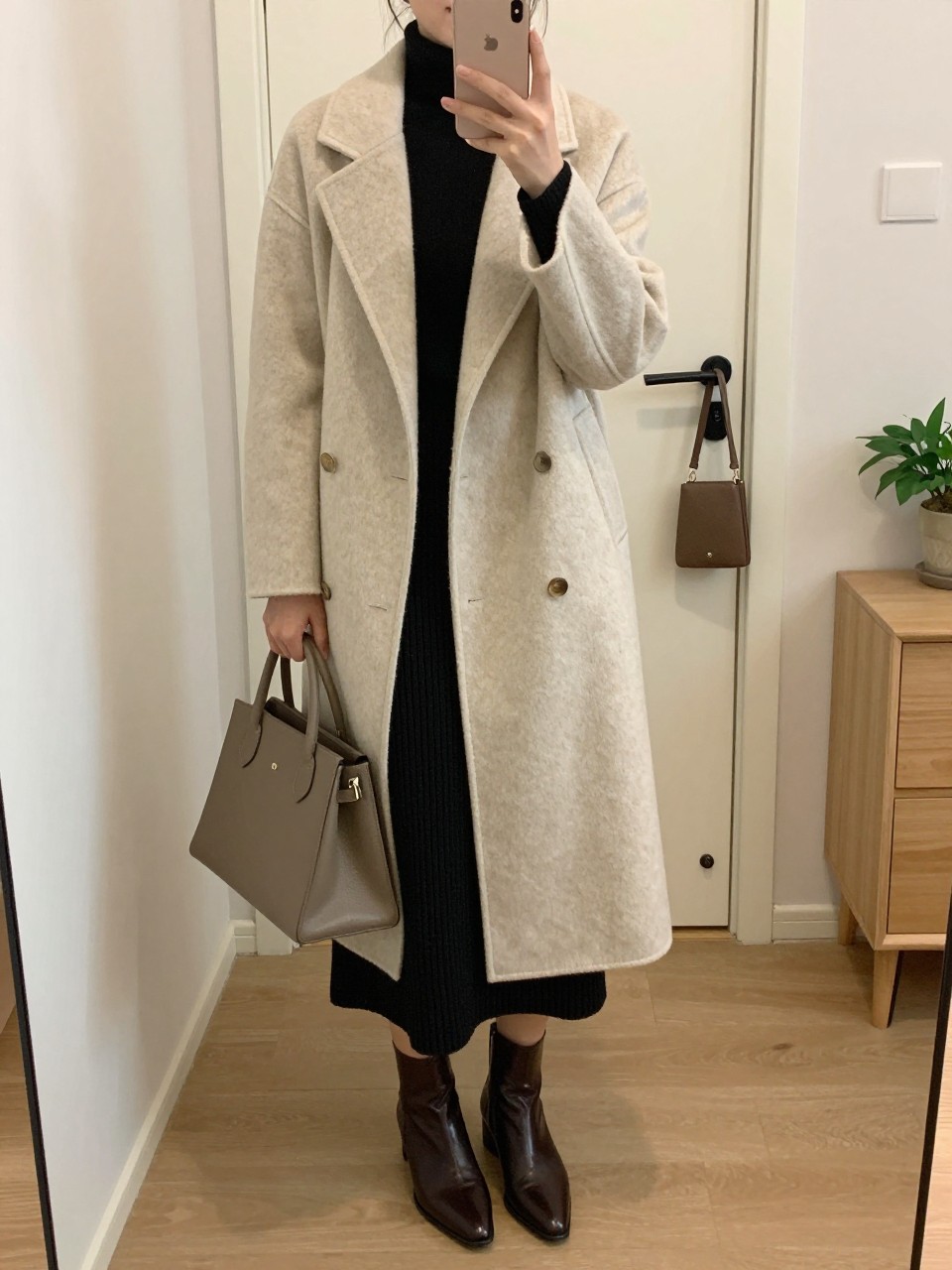 Mirror selfie of a woman wearing a long oatmeal wool coat with an attached matching scarf over a fitted black mock-neck sweater, a black ribbed-knit midi skirt, pointed dark brown leather ankle boots, and a structured taupe top-handle bag, clearly showing the exact clothing items, colors, fabrics, and accessories listed, holding a latest-model iPhone in front of a mirror, cropped at the neck so the face does not show, relaxed casual selfie pose, in a minimal dressing room with soft indoor lighting, a handbag hanging on a door handle and a small plant on a dresser, natural uncluttered setting, latest iPhone photo quality.