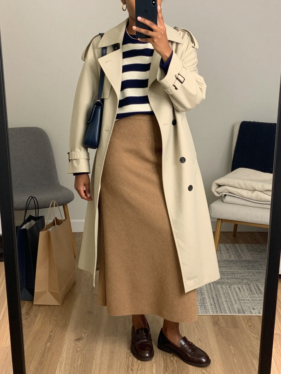 mirror selfie of a Black woman wearing the exact outfit described above, clearly showing a camel wool maxi skirt, navy and cream striped sweater, brown leather loafers, navy shoulder bag, gold huggie earrings, and a cream trench coat worn open, holding a phone in front of a mirror, cropped at NECK so the face does not show, with a relaxed casual selfie pose, in a stylish city apartment, soft indoor lighting, with shopping bags beside a chair and folded blanket on a chair, latest iPhone photo quality.