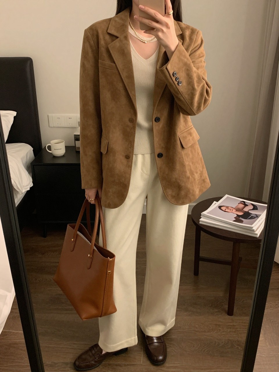 mirror selfie of a woman wearing the exact outfit described above, clearly showing a camel suede blazer layered over an ivory fitted knit top, ivory wide-leg pants, chocolate brown loafers, a brown structured tote bag, and small gold stud earrings, holding a phone in front of a mirror, cropped at NECK so the face does not show, with a relaxed casual selfie pose, in a stylish city apartment, warm moody lighting, with a coffee mug on a bedside table and a stack of fashion magazines on a side table, latest iPhone photo quality.