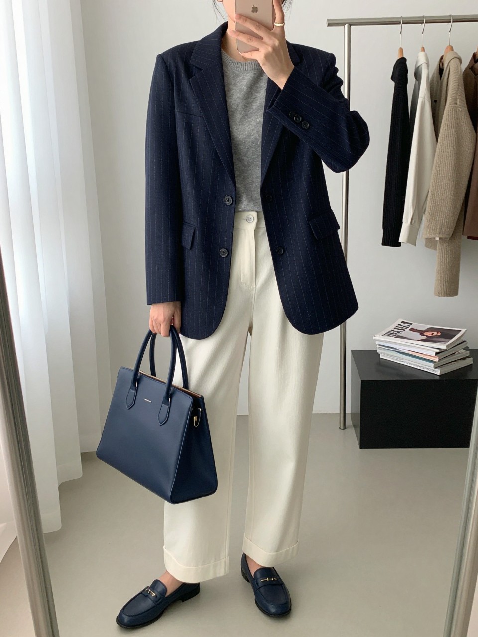 mirror selfie of a woman wearing a navy pinstripe blazer, soft gray fine-knit crewneck, ivory wide-leg trousers, navy loafers, and a navy leather satchel, clearly showing the same clothing items, colors, fabrics, and accessories listed in the Outfit Pieces section, holding a phone in front of a mirror, cropped at NECK so the face does not show, with a relaxed casual selfie pose, in a fashion studio mirror corner, natural window light, including a clothing rack with garments behind and a stack of fashion magazines on a side table, latest iPhone photo quality.