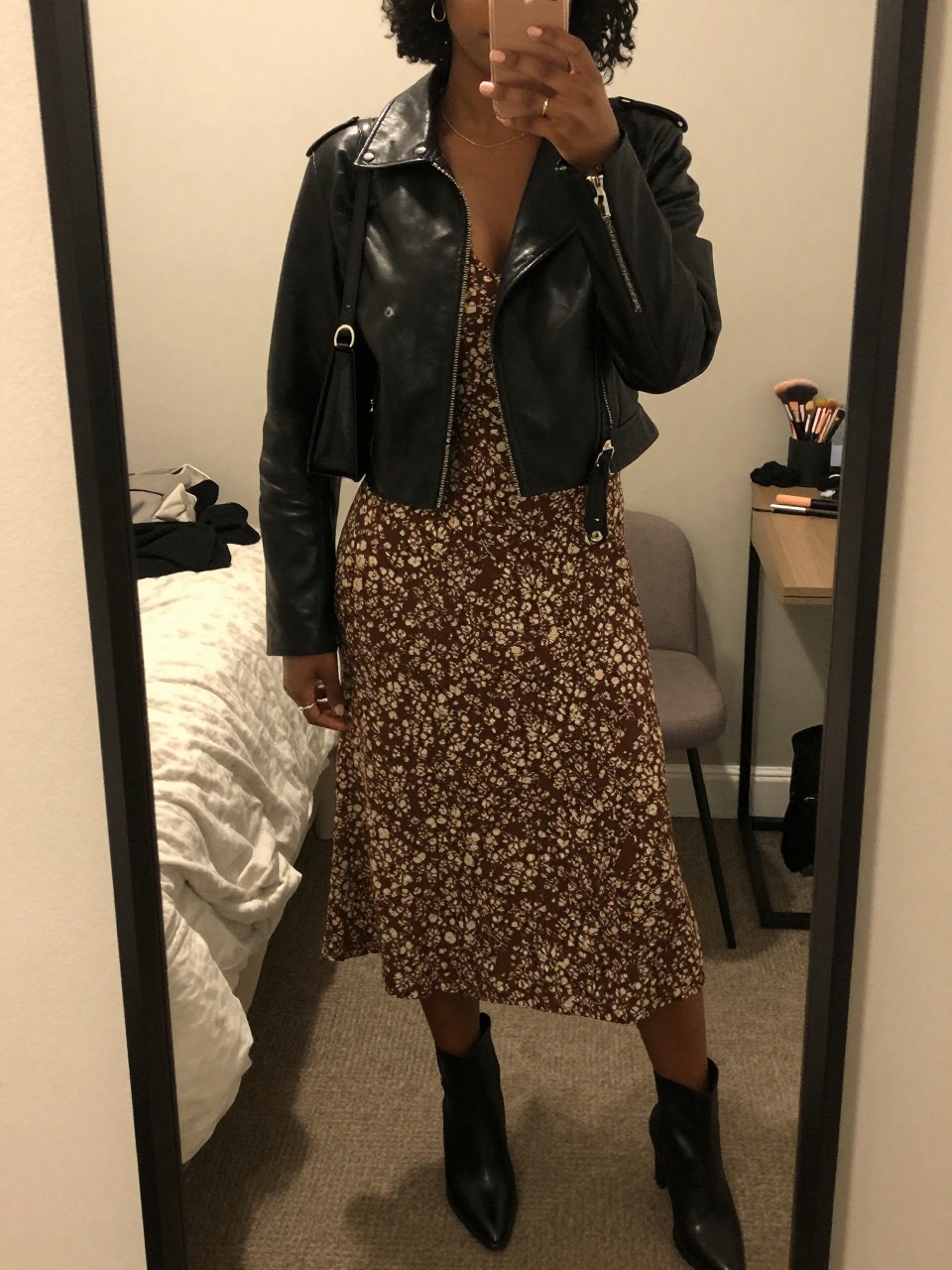 mirror selfie of a Black woman wearing a cropped black faux leather jacket, brown and cream floral midi dress, black ankle boots, a small black shoulder bag, and delicate gold earrings, clearly showing the same clothing items, colors, fabrics, and accessories listed in the Outfit Pieces section, holding a phone in front of a mirror, cropped at NECK so the face does not show, with a relaxed casual selfie pose, in a cozy bedroom mirror setup, warm moody lighting, including a folded blanket on a chair and a makeup brush on the counter, latest iPhone photo quality.