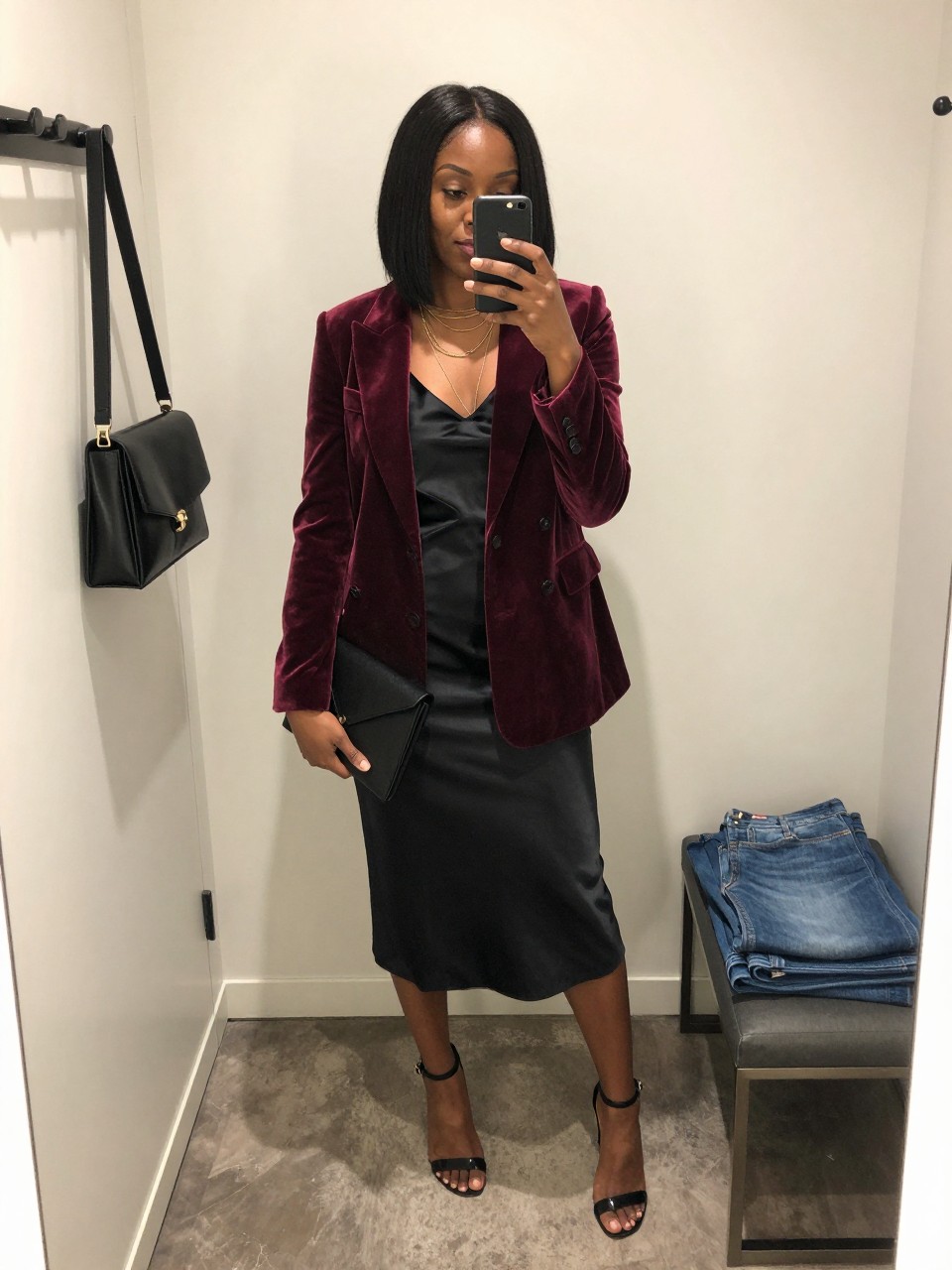 mirror selfie of a Black woman wearing the exact outfit described above, clearly showing a wine red velvet blazer layered over a black satin slip dress, black ankle-strap heels, a black envelope clutch, and gold layered necklaces, holding a phone in front of a mirror, cropped at NECK so the face does not show, with a relaxed casual selfie pose, in a minimal dressing room, soft indoor lighting, with a handbag hanging on a door handle and folded denim jeans on a bench, latest iPhone photo quality.